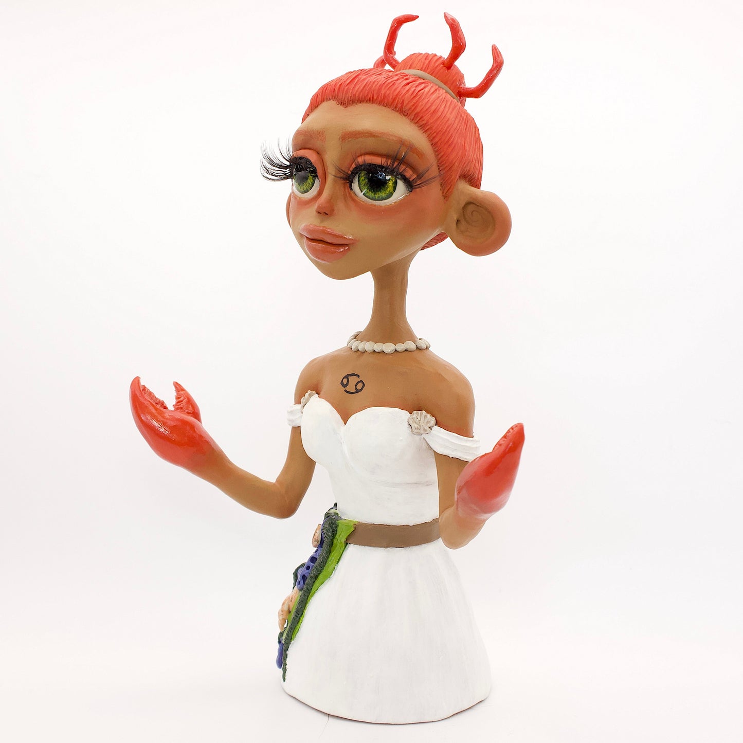 A polymer clay sculpture of the Cancer zodiac personified as a woman, featuring a seaweed belt, crab claws for hands and crab legs in her hair, she is wearing a white dress decorated with sea shells.