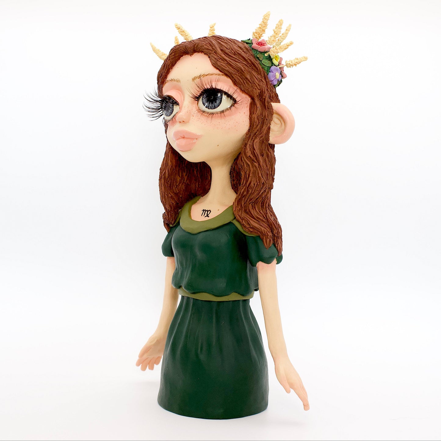 A polymer clay sculpture of the Virgo zodiac personified as a woman, featuring brown flowing hair, a forest green dress, and flowers and wheat in her hair.