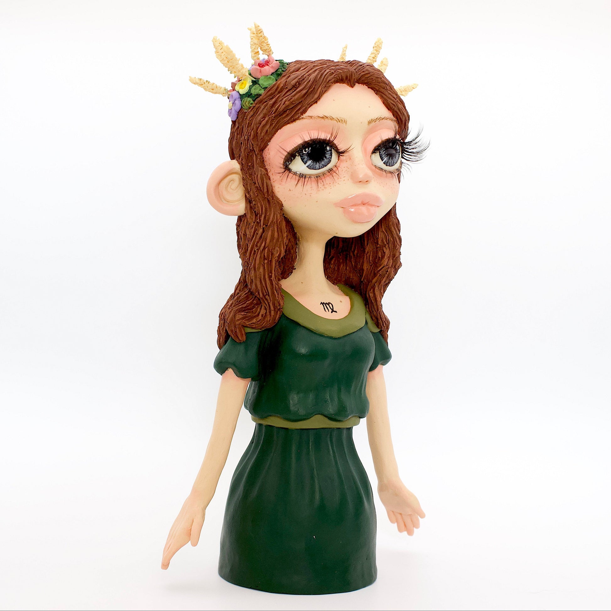 A polymer clay sculpture of the Virgo zodiac personified as a woman, featuring brown flowing hair, a forest green dress, and flowers and wheat in her hair.