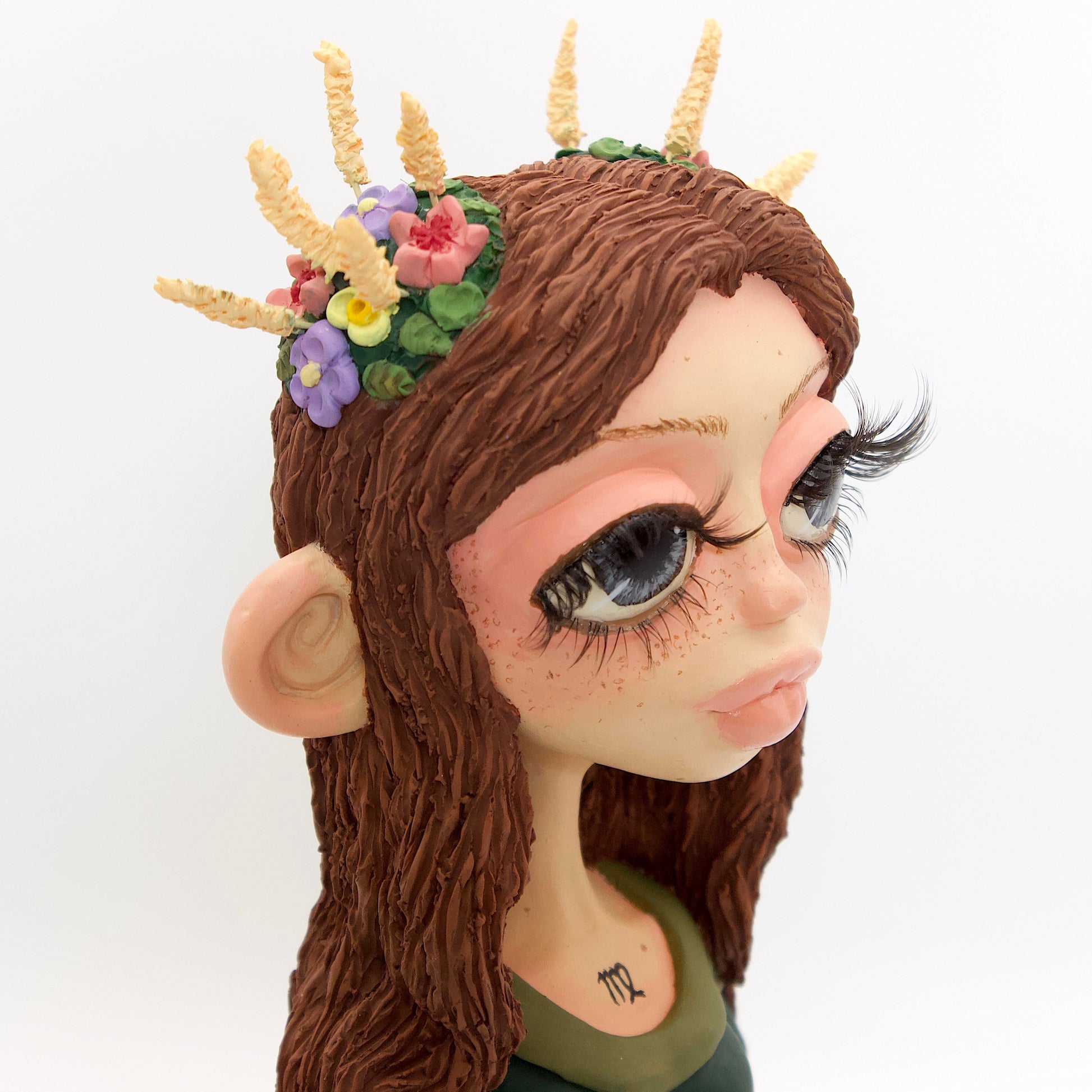 A polymer clay sculpture of the Virgo zodiac personified as a woman, featuring brown flowing hair, a forest green dress, and flowers and wheat in her hair.